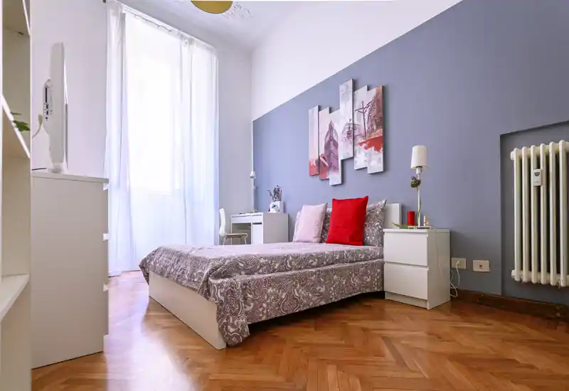 Bright, stylish bedroom with a double bed, soft linens, decorative cushions and a desk by the window. Natural light fills the room and the parquet floor and wall art add warmth.