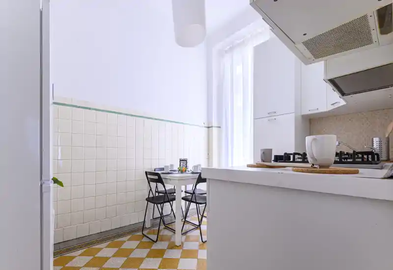 Bright, compact kitchen with a small dining table, gas stove and white cabinetry; airy window light and a warm patterned floor create a welcoming feel.