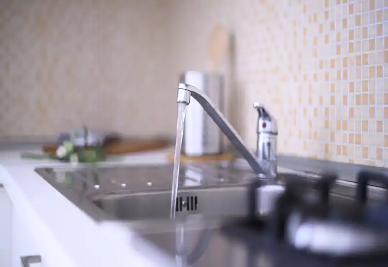 Close-up of a modern kitchen sink with a running faucet, stainless steel basin and warm mosaic tile backsplash; bright, tidy and suitable for listing photos.