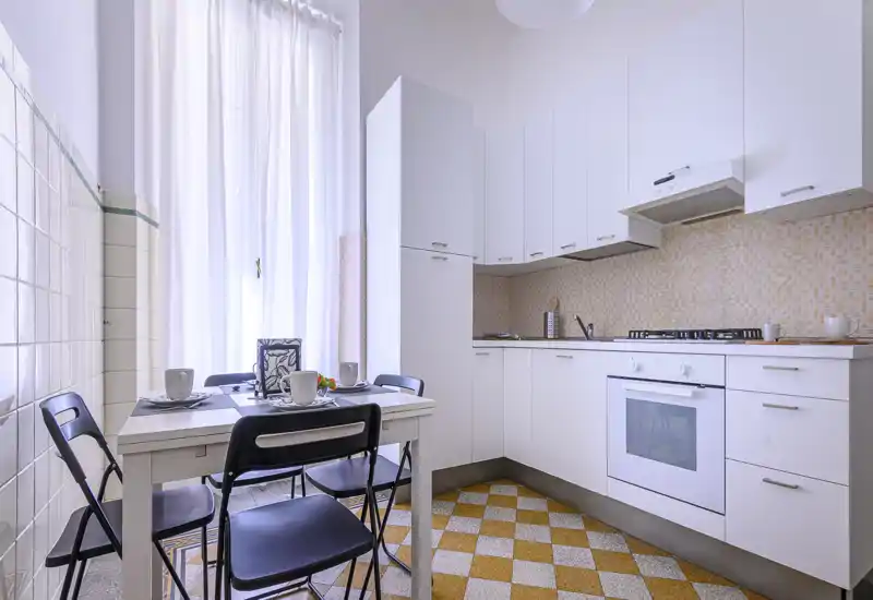 Bright, modern kitchen with white cabinets, a gas hob and built-in oven, plus a small dining table set by a tall window that fills the room with natural light.