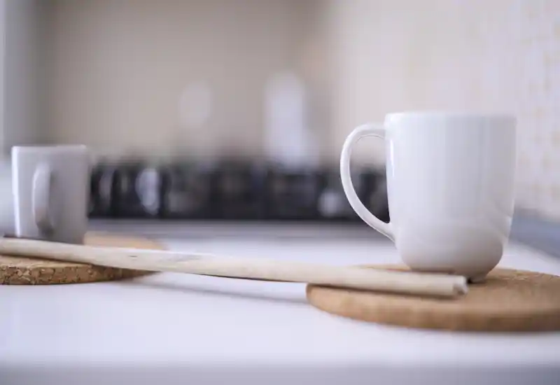 Close-up of a kitchen countertop with white mugs on cork coasters and a softly blurred stove in the background, creating a warm, minimal tabletop composition.