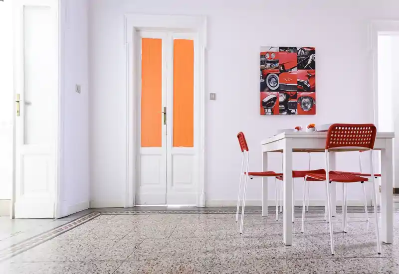 Bright, minimalist dining area with a white table and red chairs set against pale walls and decorative automotive artwork; terrazzo floor and tall doors add character and light.