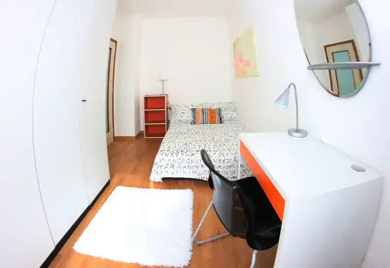 Bright, compact bedroom with a double bed, bedside lamp, small shelving and a white desk — modern, minimal and well lit.