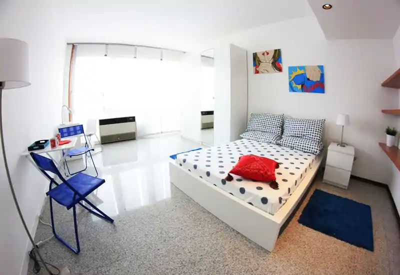 Dormitorio moderno y luminoso con cama blanca, ropa de cama con lunares y cuadros coloridos. Grandes ventanales aportan luz natural; la habitación está bien preparada para el anuncio.