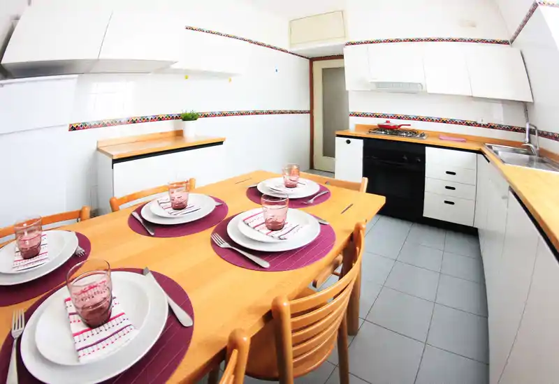 Bright, tidy kitchen with a wooden dining table set for four, white cabinets, a built-in oven and a stainless sink — ideal for casual meals.