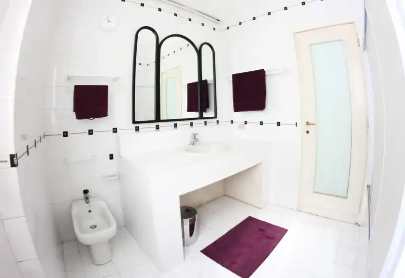 Baño luminoso todo en blanco con lavabo integrado en mueble alicatado, bidé, gran espejo decorativo y toallas/alfombra burdeos; espacio limpio y bien iluminado apto para anuncios.