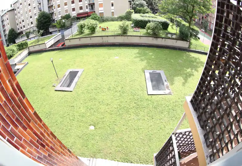 Patio comunitario luminoso con césped bien cuidado, bordeado por setos y bloques de apartamentos: ideal para relajación al aire libre y juegos infantiles.