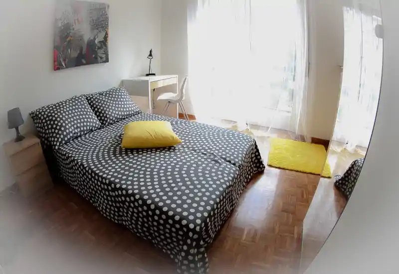 Dormitorio luminoso y moderno con colcha de lunares, cojín amarillo y un pequeño escritorio junto a la ventana que aporta mucha luz natural.