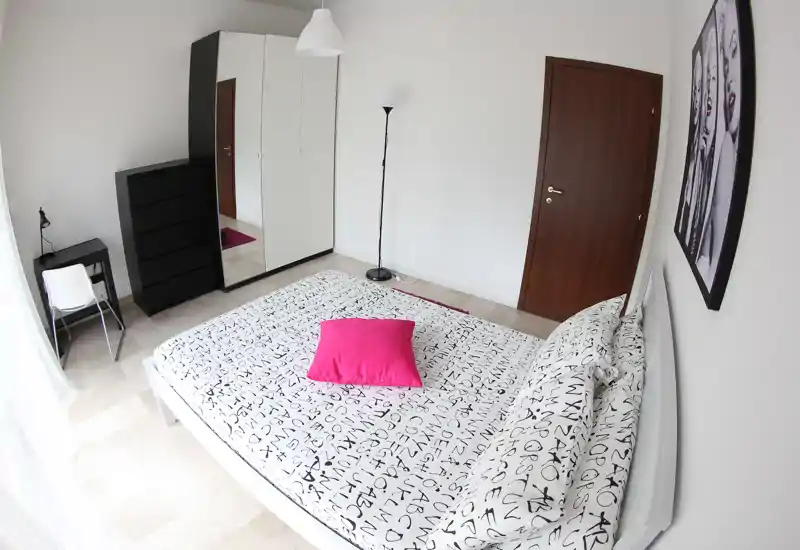 Dormitorio moderno y luminoso con cama doble con ropa de cama estampada y cojín rosa intenso, armario con espejo y mobiliario minimalista.