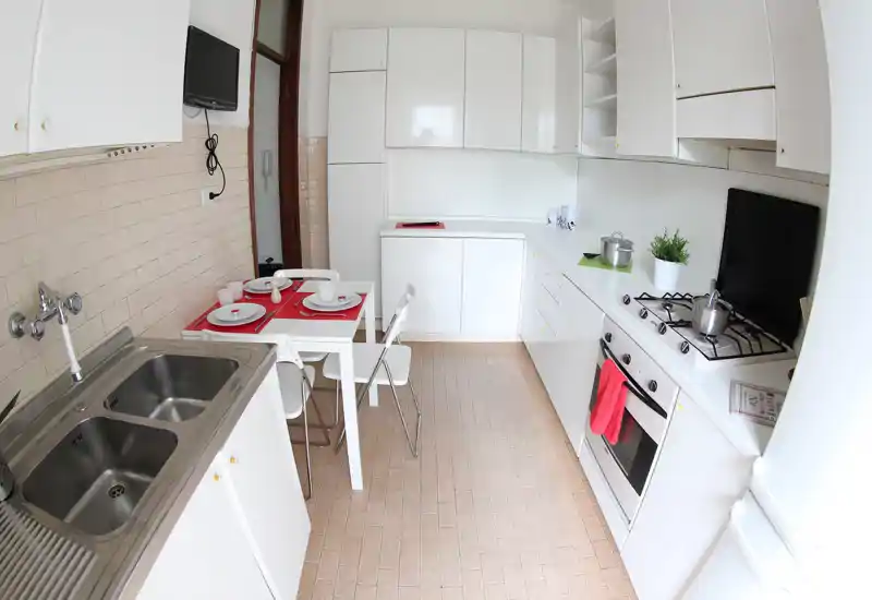 Cocina compacta y luminosa con armarios blancos, cocina de gas, horno empotrado y una pequeña mesa para dos — ordenada y moderna para un piso urbano.