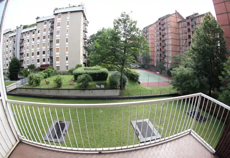 Vistas desde el balcón a un patio comunitario con césped, árboles y pista deportiva; perspectiva gran angular que muestra bien el espacio exterior.
