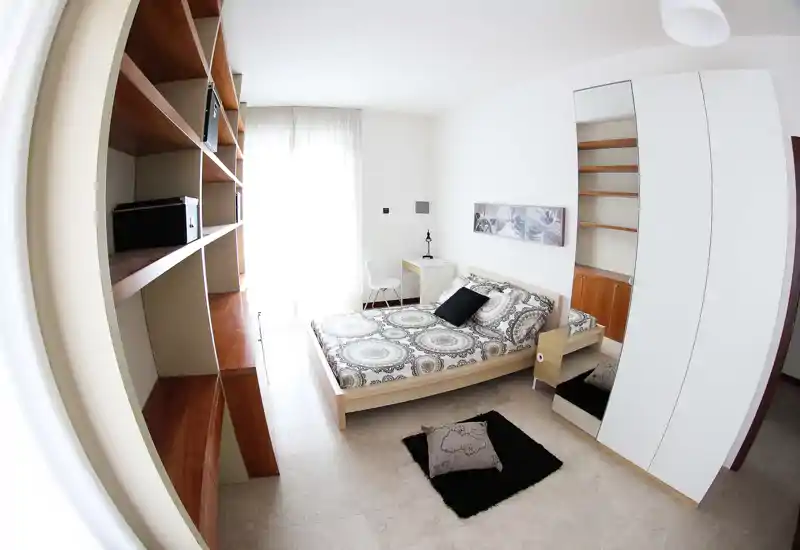 Bright, modern bedroom with a double bed, patterned bedding and a mirrored wardrobe. Large window with sheer curtains fills the room with natural light and a built-in shelving unit adds storage.