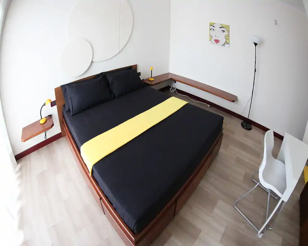 Dormitorio moderno y luminoso con cama doble grande vestida en negro y una banda amarilla, mesitas minimalistas y luz natural suave.