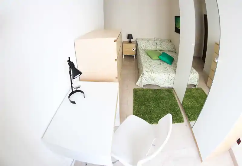 Compact, bright bedroom with a double bed dressed in green patterned linens, matching cushions and a green rug. A white desk with a lamp and a full-length mirrored wardrobe create a neat, functional layout ideal for a single occupant or short stays.