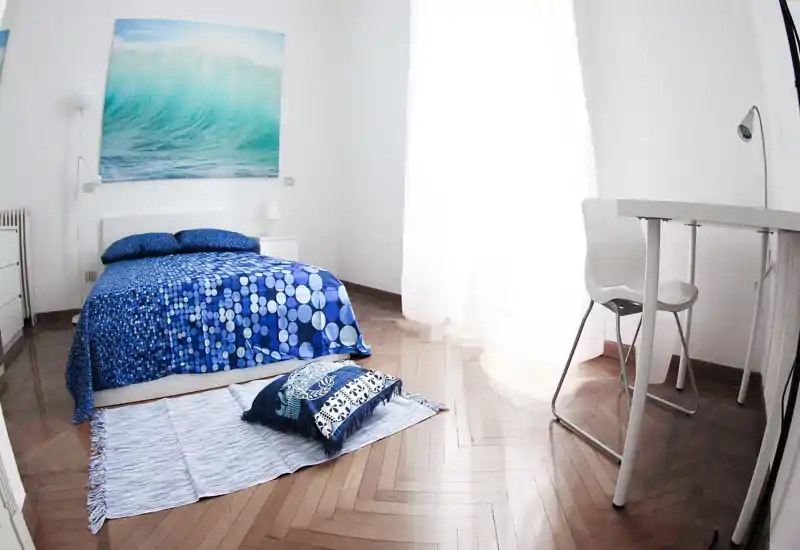 Camera da letto luminosa e moderna con letto matrimoniale con biancheria blu a fantasia, cuscini decorativi e piccolo tappeto; ampia finestra con molta luce naturale e un'area scrivania minimalista sul lato.