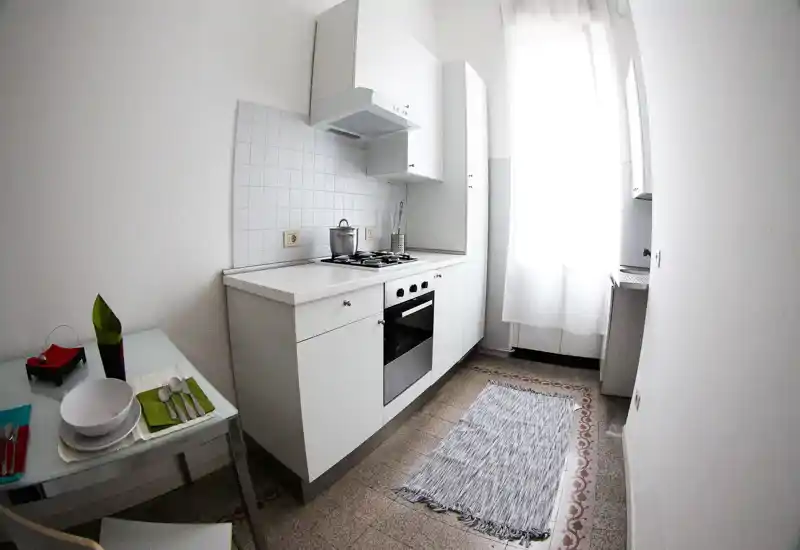 Cucina compatta e luminosa con mobili bianchi, piano cottura a gas e forno integrato; piccolo angolo pranzo con tovagliette e tappeto decorativo vicino a una finestra soleggiata.