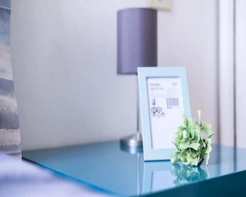 Close-up of a bedside table in a bedroom featuring a turquoise glossy surface, framed photo, decorative faux flowers and a lamp — bright, styled detail shot ideal for listing highlights.