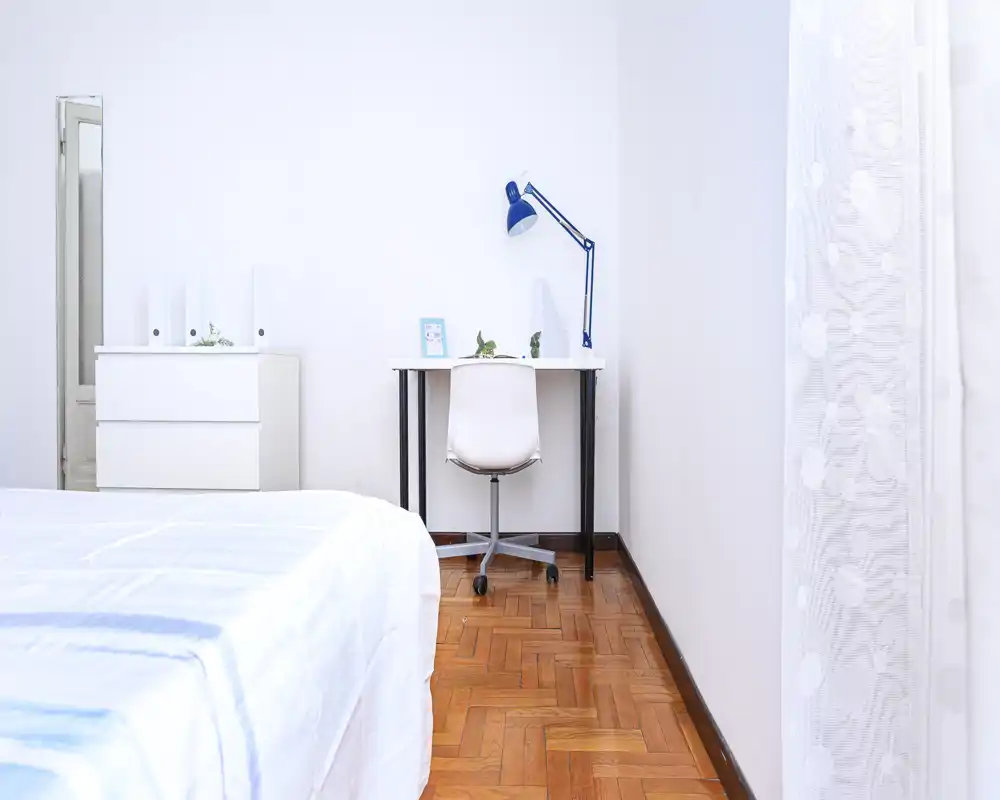 Bright, minimalist bedroom with a visible bed in the foreground, a small desk with a chair and lamp against the wall, and a dresser with a mirror — clean, airy and uncluttered.