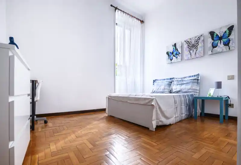 Bright, airy bedroom with a double bed dressed in light linens, wooden parquet flooring and butterfly wall art; natural light floods through a tall sheer-curtained window.
