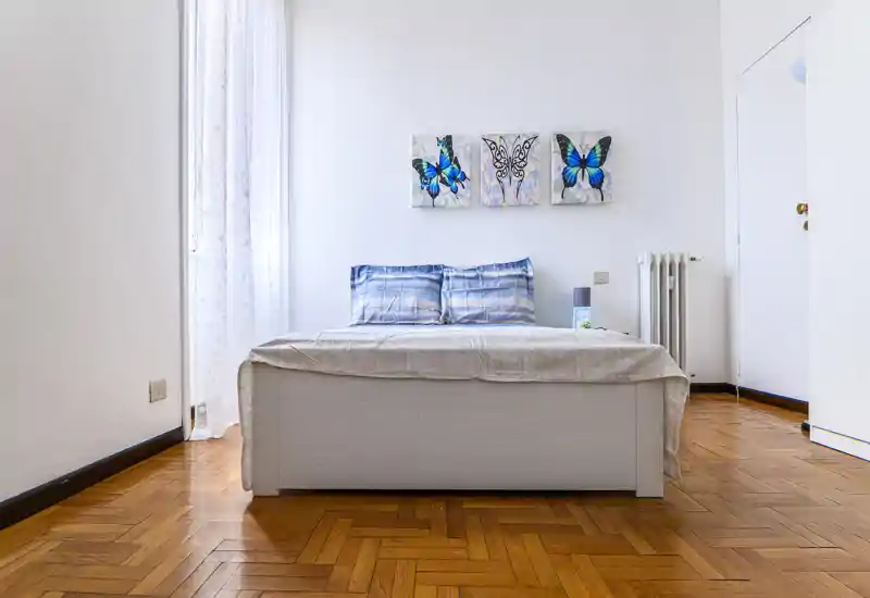 Bright, minimalist bedroom with a centrally placed double bed, blue patterned pillows, light linens and butterfly wall art; hardwood floors and natural light from a tall window create an airy feel.