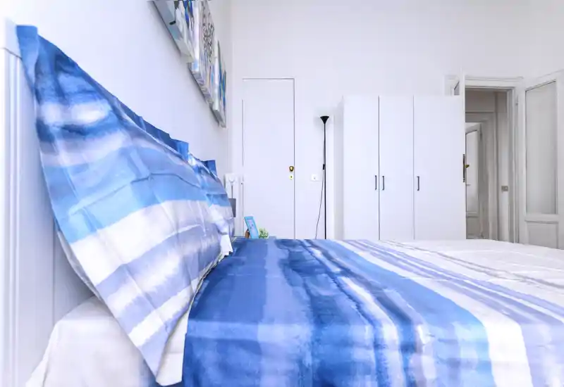 Bright modern bedroom with a large bed dressed in blue-striped linens, a white wardrobe and minimalist décor — well lit and clean, ideal for rental listings.