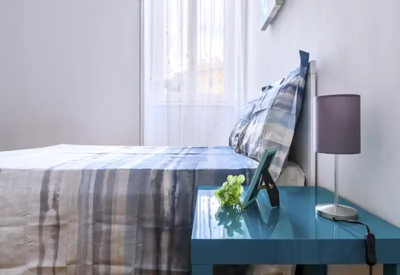 Bright, neatly made bedroom with a patterned duvet, bedside table with lamp and small decor, and soft natural light from the window.