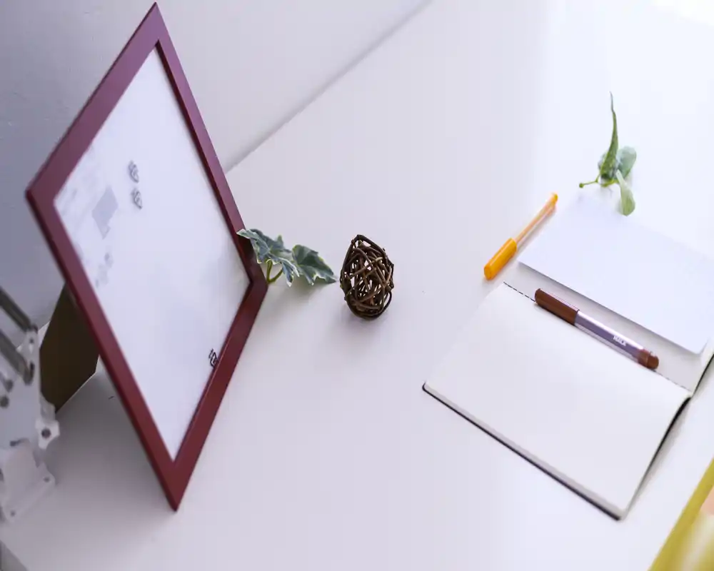 Close-up of a tidy living area surface with a notebook, pens, a decorative picture frame and small greenery — bright, minimal and neatly styled.