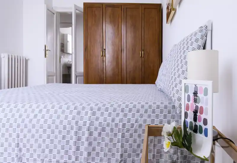 Bright, well-staged double bedroom with patterned linens, a wooden wardrobe and a bedside table with lamp and flowers — clean, inviting space ideal for holiday stays.