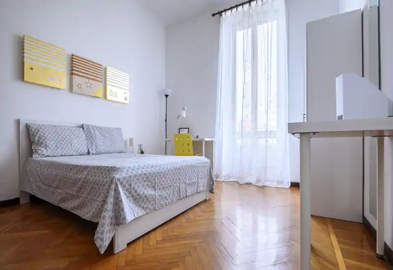 Bright, modern bedroom with a double bed, patterned bedding and a large window with sheer curtains letting in abundant natural light; a small desk and chair sit by the window, and warm parquet flooring completes the calm, airy space.
