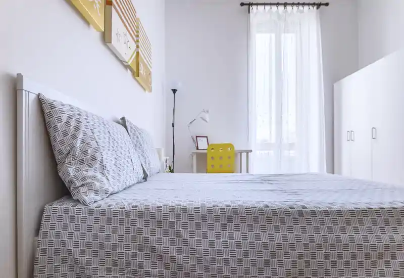 Bright, minimal bedroom featuring a neatly made double bed with patterned linens, a desk with a yellow chair by the window, and a tall wardrobe — airy, modern and well-lit.