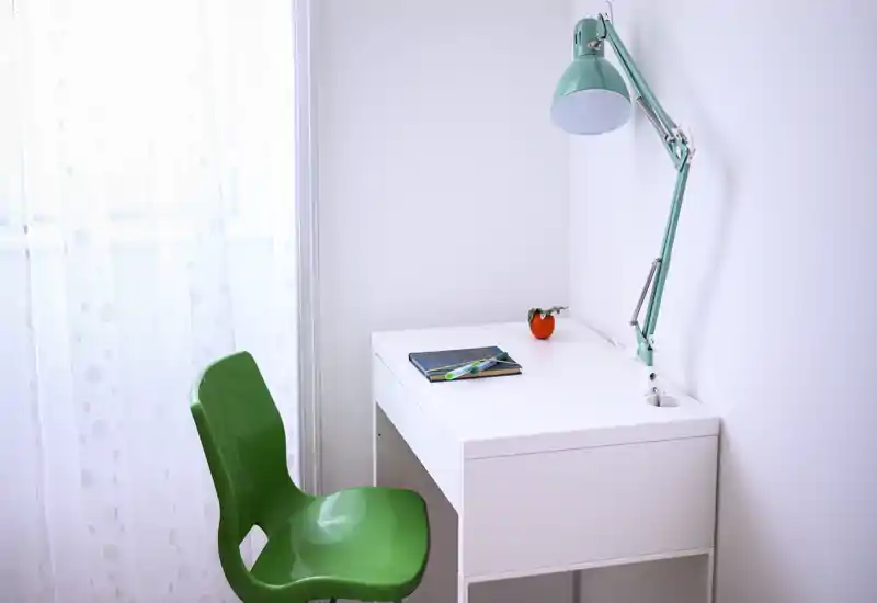 Angolo studio luminoso e minimale con scrivania bianca, sedia verde e lampada da tavolo turchese regolabile vicino a una finestra con tenda leggera — ideale per lavorare o studiare.