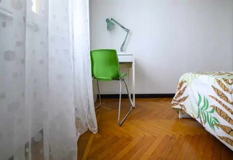 Angolo camera luminoso e minimale con una scrivania, sedia verde e letto con copriletto a fantasia; ideale per una persona che cerca uno spazio moderno e ordinato.