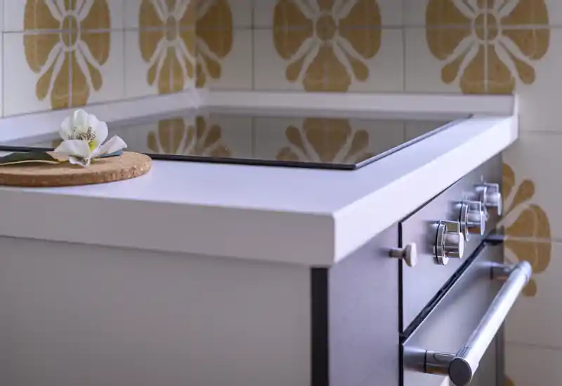 Close-up of a modern kitchen cooktop and oven controls, with a decorative flower on the countertop and patterned tile backsplash.