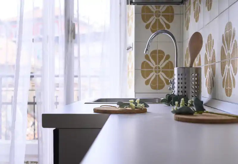 Bright, styled kitchen countertop with sink next to a sheer-windowed balcony; decorative placemats and small plant accents add warmth.