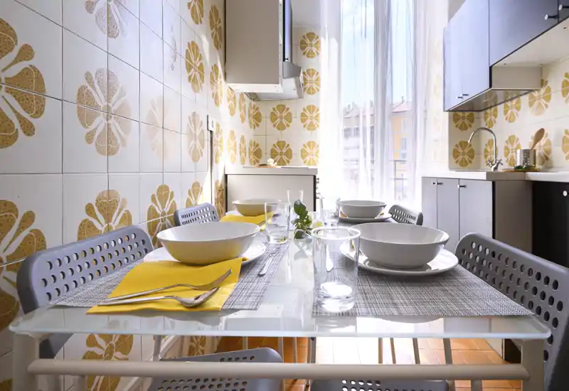 Cucina luminosa e arredatta con tavolo in vetro per quattro, piastrelle decorate e luce naturale da una grande finestra — perfetta per mostrare una cucina compatta in appartamento.