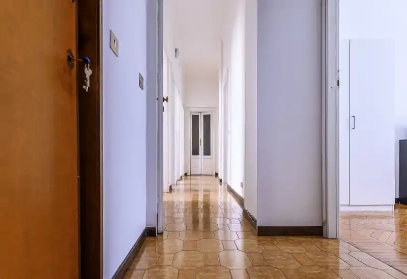 Bright, long apartment corridor with glossy patterned floor and white walls leading to a glass-paneled door; clean, well-lit entrance hallway.