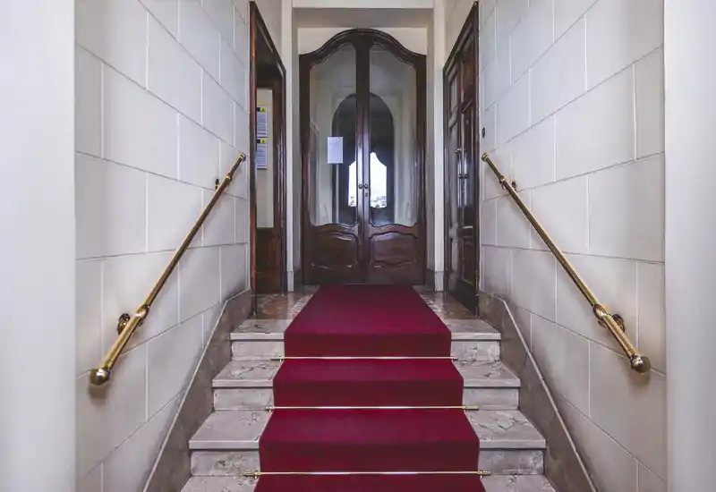 Elegant building entrance with a short marble staircase covered by a red runner, brass handrails and double wooden doors — well-lit, symmetrical and welcoming.