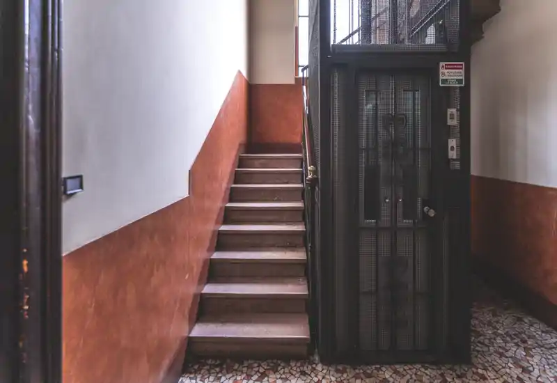 Bright, well-maintained building entrance with a tiled floor, a narrow staircase and a vintage elevator with decorative metal grille—warm terracotta wall paint adds character.