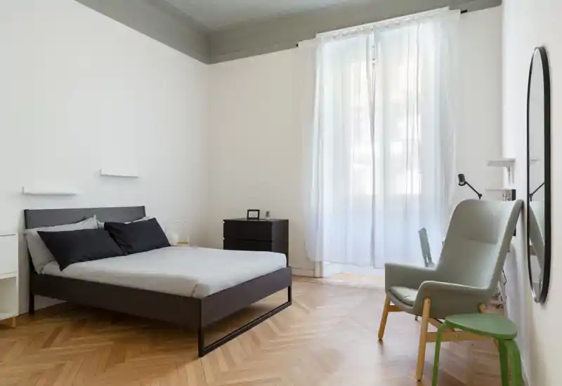 Camera da letto luminosa e minimalista con letto in legno scuro, biancheria neutra e una poltrona comoda vicino alla finestra. Ampia finestra con tende leggere che diffondono luce naturale; pavimento in parquet e arredi semplici creano un’atmosfera serena ideale per annunci.