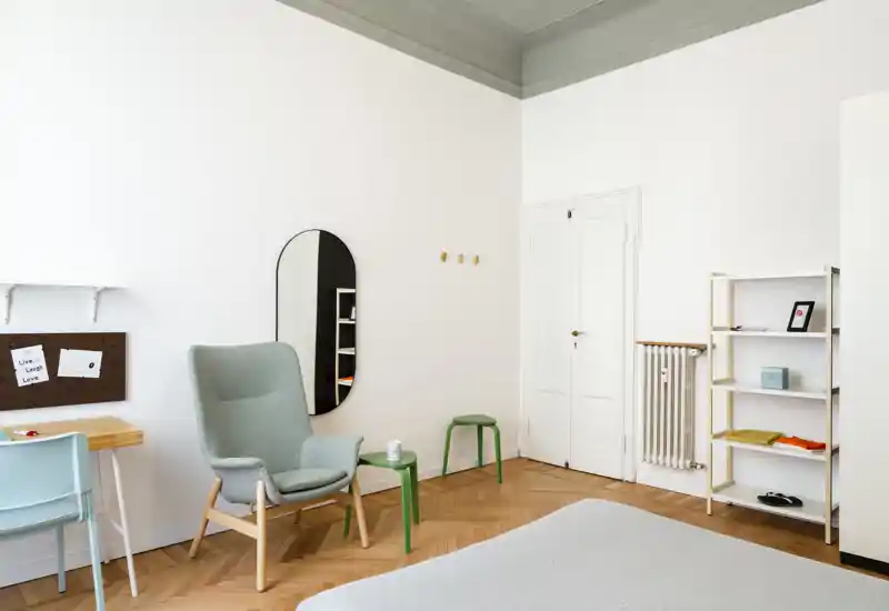 Camera da letto luminosa e minimal con letto in primo piano, una comoda poltrona, tavolini e una libreria; ambiente arioso e tranquillo ideale per un annuncio di affitto.