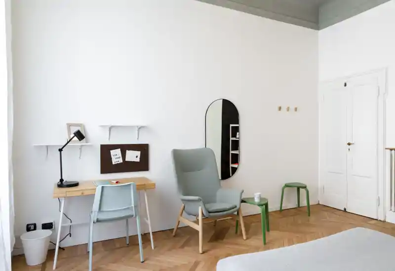 Angolo camera da letto luminoso e minimale con una poltrona grigio chiaro, una piccola scrivania in legno e il letto parzialmente visibile sul lato; stile scandinavo moderno e pavimento a spina di pesce in legno.