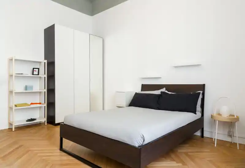 Camera da letto luminosa e minimal con letto in legno scuro, biancheria neutra e un armadio semplice: perfetta per annunci che puntano su uno stile moderno e pulito.