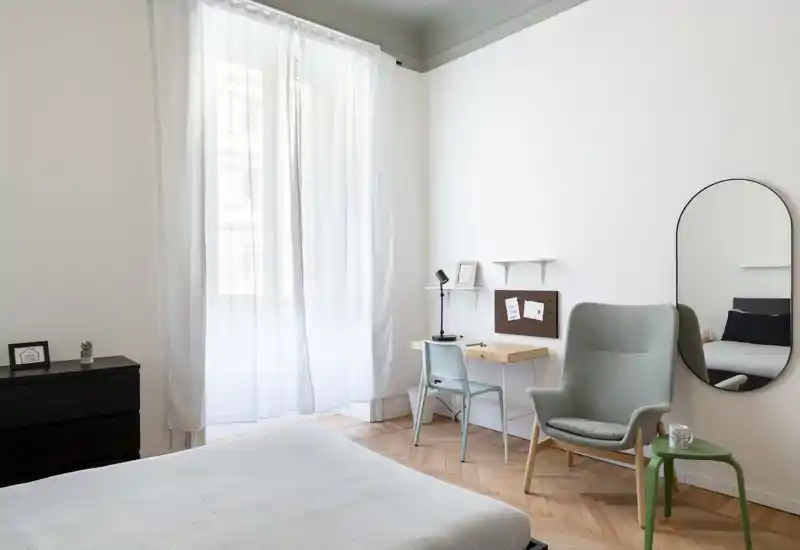Camera da letto luminosa e minimalista con grande finestra, letto in primo piano, una scrivania e una poltrona — ideale per un affitto tranquillo e arioso.