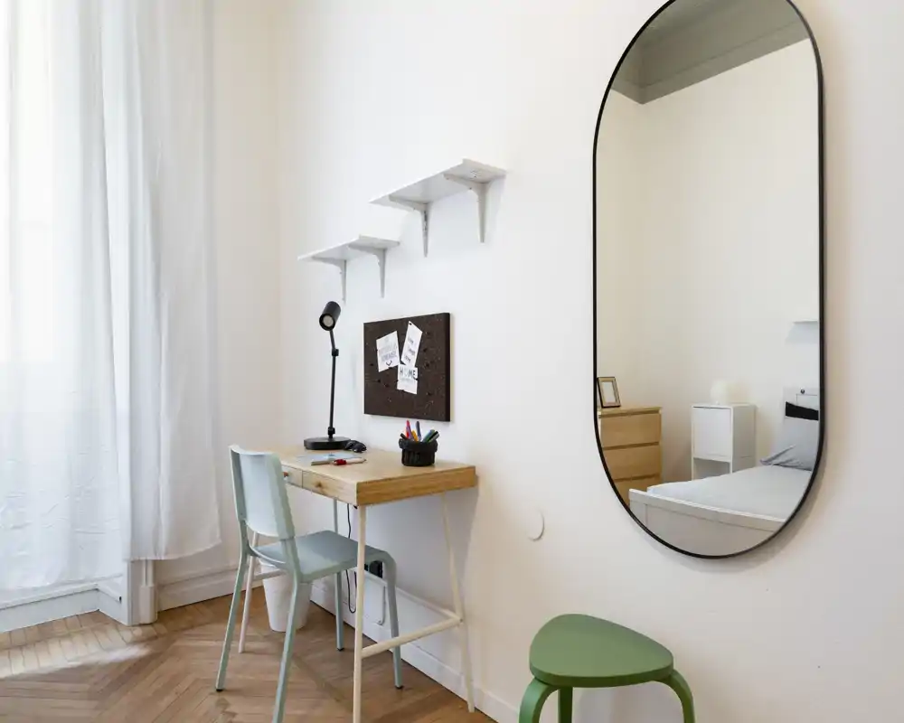 Angolo luminoso di camera da letto con scrivania e uno specchio che riflette parzialmente il letto; luce naturale morbida e arredi puliti in stile scandinavo.