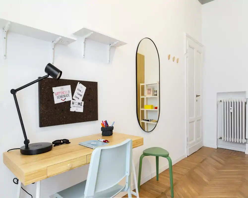 Angolo studio luminoso e minimal con scrivania in legno, lampada da tavolo, sedia e bacheca in sughero — uno spazio di lavoro ordinato integrato nell'ambiente.