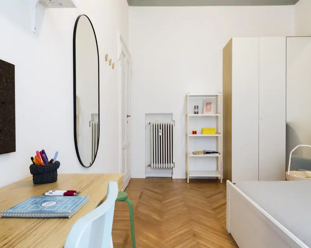 Camera da letto luminosa e moderna con un semplice letto, armadio e una scrivania in legno chiaro. Linee pulite, pavimento a spina di pesce caldo e dettagli decorativi minimali rendono la stanza accogliente e funzionale.