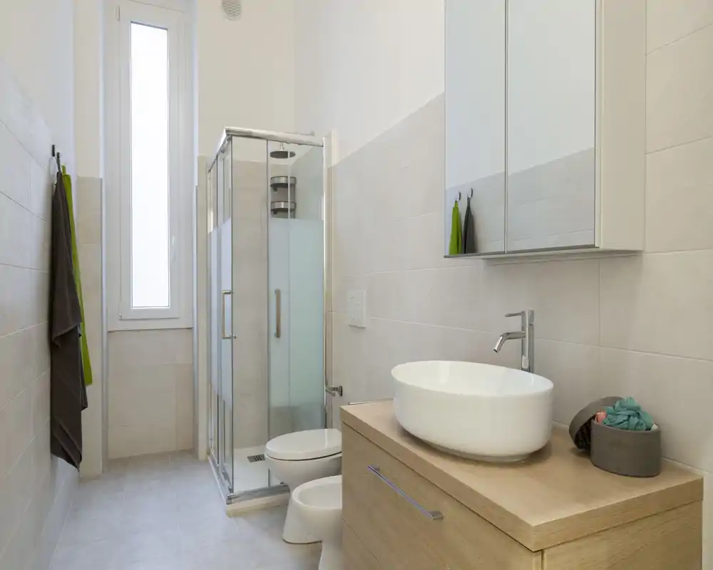 Bagno moderno e luminoso con cabina doccia in vetro, WC a parete e mobile lavabo effetto legno con lavabo a bacinella bianco. Piastrelle neutre e una finestra alta satinata donano luce naturale e un’atmosfera pulita.