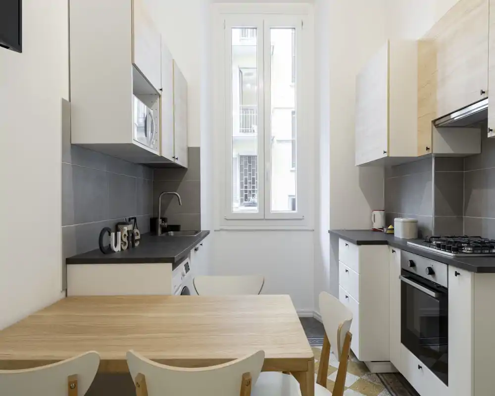 Cucina moderna e luminosa con tavolo in legno, forno incassato e piano cottura a gas, ampia finestra che garantisce luce naturale — perfetta per un appartamento compatto.