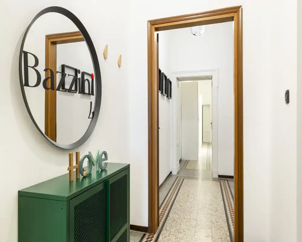 Ingresso luminoso e accogliente con specchio rotondo decorativo, mobile consolle verde e corridoio piastrellato che conduce all'appartamento.
