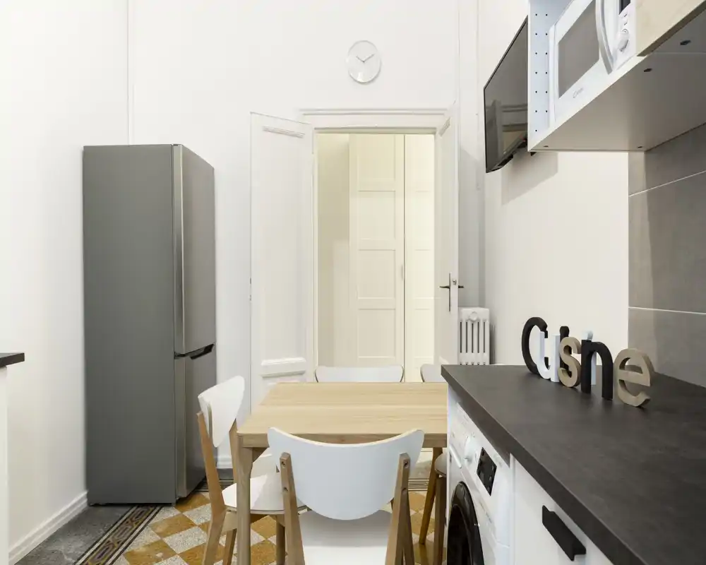 Cucina compatta e luminosa con tavolo in legno, elettrodomestici moderni e pareti bianche, perfetta per un appartamento di piccole dimensioni.